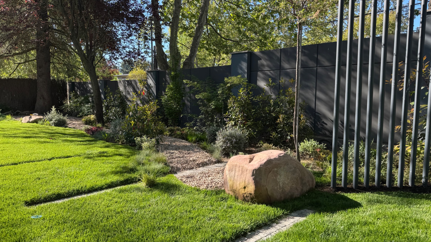 Lawn with a large boulder, gravel path and vertical metal screen beneath mature trees